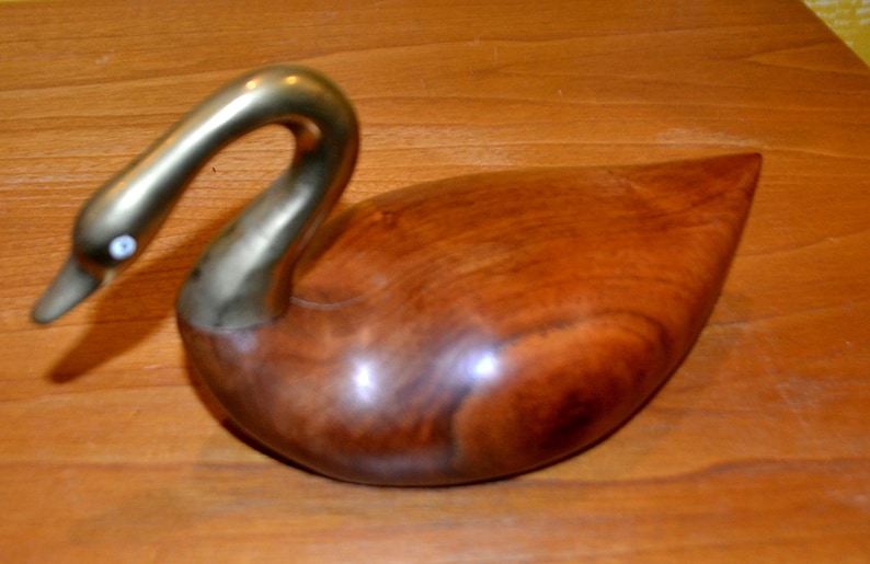 May include: A wooden swan sculpture with a brass neck and head. The swan has a white eye and is resting on a wooden surface.