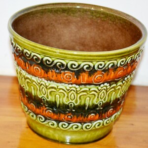 Op de afbeelding: Een keramische plantenbak met een glanzende afwerking. De plantenbak heeft banden van oranje, groene en zwarte decoratieve patronen. De binnenkant is donkerbruin van kleur en de buitenkant heeft een groene basis en rand.