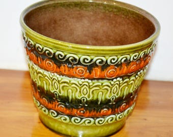 May include: A ceramic planter with a glossy finish. The planter features bands of orange, green, and black decorative patterns. The interior is a dark brown color, and the exterior has a green base and rim.
