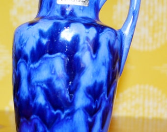May include: Blue and white ceramic vase with a swirl pattern. The vase has a handle and a label that reads "Sauerich nur die Linie".