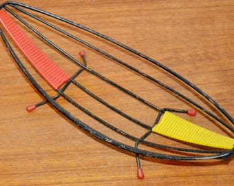 Op de afbeelding: Een vintage zwarte metalen trivet met een uniek ontwerp. Het heeft gebogen metalen staven die een ovale vorm vormen, met rode en gele accenten. De trivet heeft kleine rode voetjes.