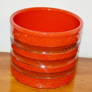 Könnte beinhalten: Orangener Keramiktopf mit strukturierter Oberfläche und vier braunen Bändern um die Mitte.