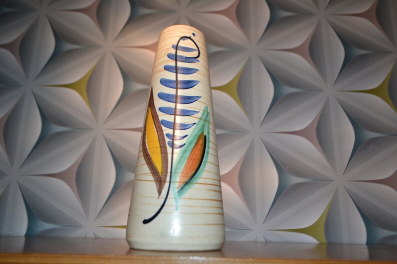 May include: A white ceramic vase with a tall, slender shape and a geometric pattern of brown, blue, yellow, and green leaves.