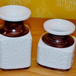 May include: Two white ceramic vases with brown accents and a floral design. The vases are square shaped with a wide opening at the top.