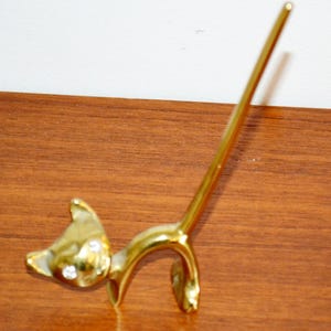 Könnte beinhalten: Goldfarbene Katzenfigur mit langem, nach oben gebogenem Schwanz. Der Kopf der Katze hat zwei klare, funkelnde Akzente. Die Figur besteht aus glänzendem Metall und ist in einer spielerischen Haltung dargestellt.