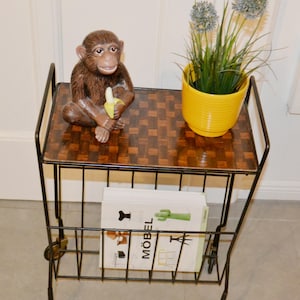 May include: A vintage black metal rolling cart with a patterned wooden top. A brown monkey figurine holding a banana sits on top. A yellow pot with greenery and a magazine with the word "MÖBEL" are also on the cart.