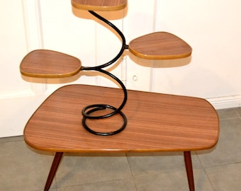 May include: A mid-century modern plant stand with a brown and black color scheme. The stand features a large, rectangular tabletop supported by three angled legs. A black, curved metal structure rises from the table, supporting three smaller, leaf-shaped shelves.