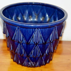 May include: A cobalt blue ceramic planter with a geometric design. The planter has a cylindrical shape with a slightly flared rim. The exterior features a repeating pattern of inverted triangles with vertical lines, creating a textured surface.