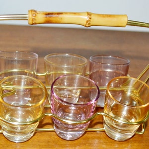 Op de afbeelding: Een gouden metalen shotglazenhouder met een bamboe handvat. De houder bevat zes shotglazen, waarvan twee helder, twee amberkleurig en twee roze zijn.