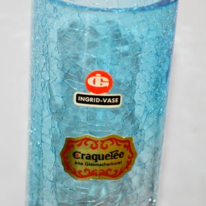 Könnte beinhalten: Eine hohe, zylindrische, hellblaue Vase mit einem Craquelé-Glaseffekt. Die Vase hat zwei Etiketten: eines mit dem Text "INGRID-VASE" und ein weiteres mit "Craquelée Alte Glasmacherkunst". Die Vase ist ein dekorativer Gegenstand.