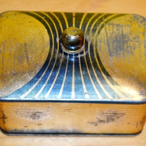 Puede incluir: Una caja de metal vintage dorada y azul con un diseño decorativo y una perilla redonda en la tapa.