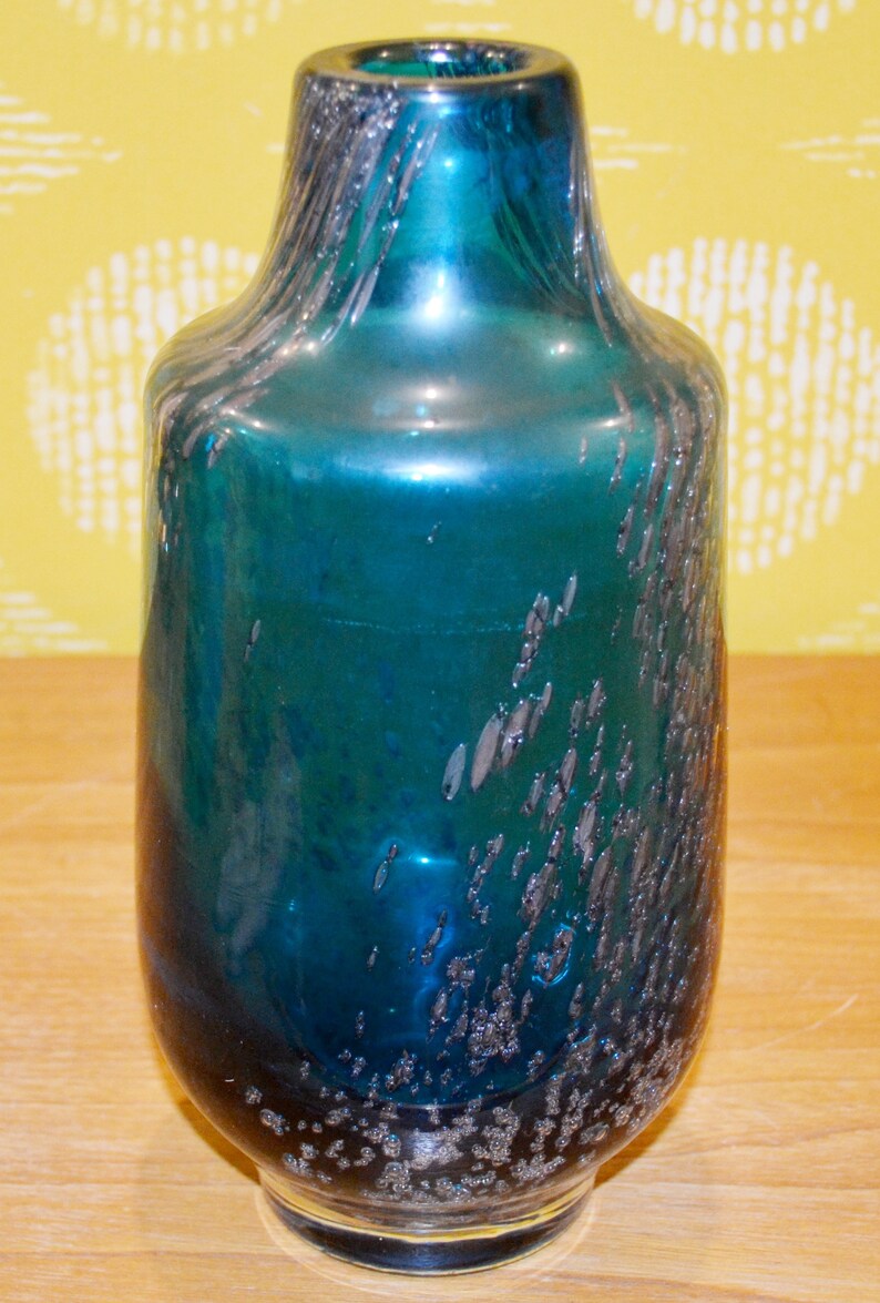 May include: A teal blue glass vase with a textured, speckled finish. The vase has a rounded body and a narrow neck.