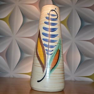 May include: A tall, cream-colored ceramic vase with a stylized leaf design in blue, green, yellow, and orange. The vase has a textured surface and a slightly flared opening.