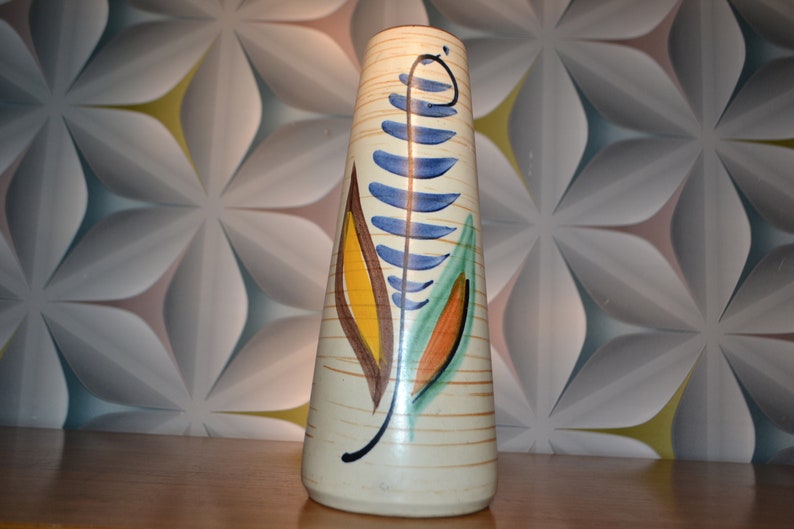 May include: A tall, cream-colored ceramic vase with a stylized leaf design in blue, green, yellow, and orange. The vase has a textured surface and a slightly flared opening.