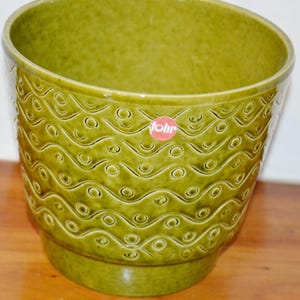 Può includere: Un vaso per piante in ceramica color verde oliva, di forma rotonda, caratterizzato da un motivo in rilievo di linee ondulate orizzontali e piccoli cerchi. Sulla superficie esterna smaltata è presente un adesivo rosso con la scritta "fohr".
