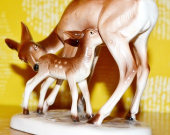 May include: A porcelain figurine of a doe and fawn. The doe is brown with white markings and the fawn is brown with white spots. The figurine is on a white base.