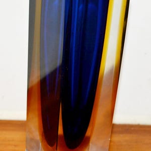 Könnte beinhalten: Eine rechteckige Glasvase mit quadratischer Öffnung. Die Vase hat einen Farbverlauf aus Blau, Gelb und Orange. Der Innenraum ist hauptsächlich blau, mit gelben und orangen Akzenten an den Seiten. Die Vase steht auf einer Holzoberfläche.