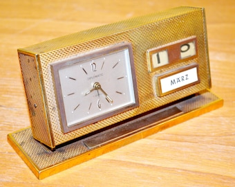May include: A gold-toned desktop clock with a calendar feature. The clock face reads "DIPLOMATIC" and the calendar shows the date "19" and the month "MARZ".