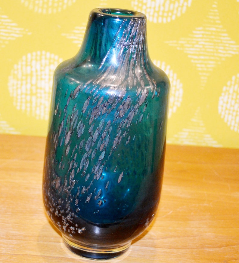 May include: A teal blue glass vase with silver flecks throughout. The vase has a round body and a narrow neck.