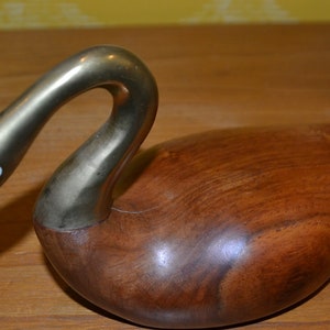 May include: A wooden swan sculpture with a brass neck and head. The swan has a white eye and is resting on a wooden surface.