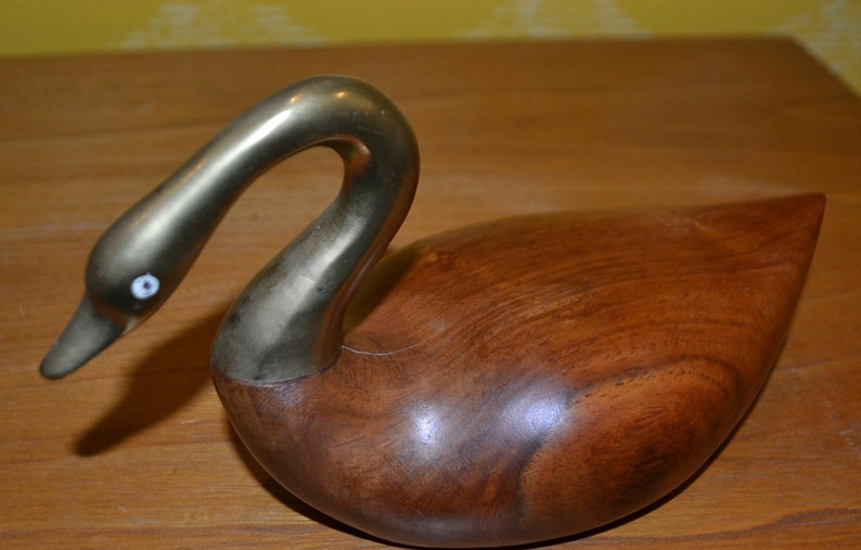 May include: A wooden swan sculpture with a brass neck and head. The swan has a white eye and is resting on a wooden surface.