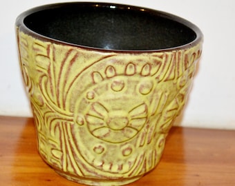 May include: A ceramic planter with a yellow-green exterior and a dark brown interior. The planter features a raised, repeating floral and geometric pattern in a darker brown color. The rim is angled, and the interior is smooth.