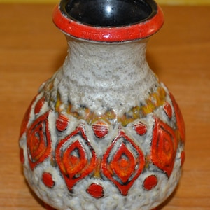 Könnte beinhalten: Eine kleine, weiße Keramikvase mit rotem Rand und einem geometrischen Muster in Orange- und Rottönen. Die Vase hat eine strukturierte Oberfläche und ein schwarzes Inneres.