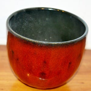 Puede incluir: Un macetero de cerámica rojo y negro con forma redondeada. El macetero tiene un esmalte verde oscuro en el interior.