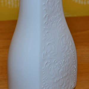 May include: A white ceramic vase with an embossed floral design. The vase has a rectangular shape with a wide base and a narrow neck.