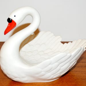 May include: A white ceramic swan figurine with a red beak and black eyes. The swan has a curved neck and detailed wing feathers. The figurine is likely a decorative item, possibly a dish or a small container.
