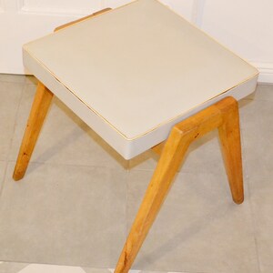 May include: A small, square, white upholstered stool with a light wood frame and four angled legs.
