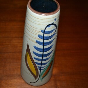 May include: A white ceramic vase with a blue, green, yellow, and brown floral design. The vase has a cylindrical shape and a slightly flared opening.