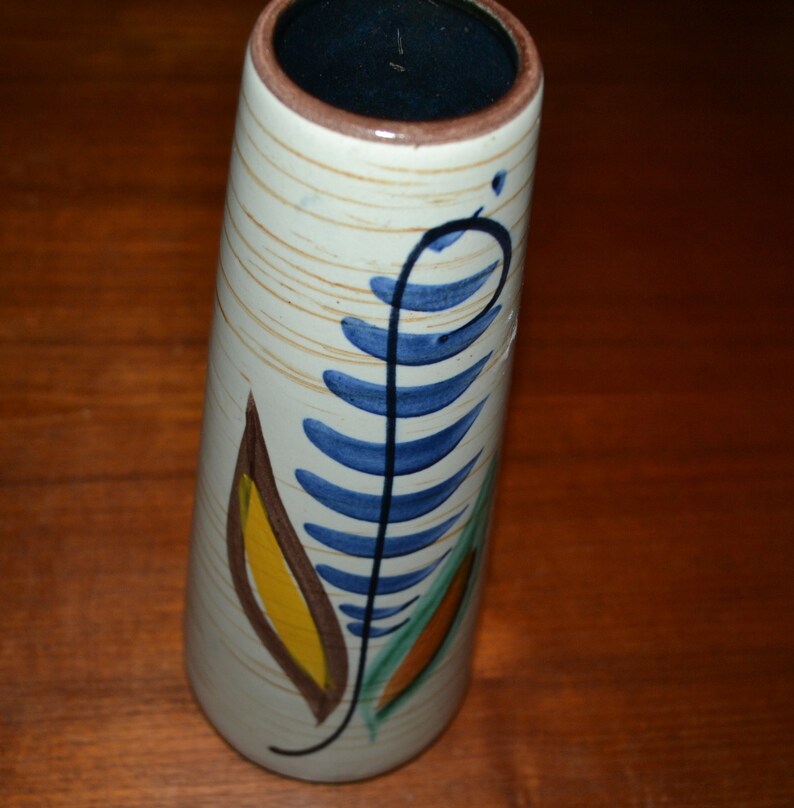 May include: A white ceramic vase with a blue, green, yellow, and brown floral design. The vase has a cylindrical shape and a slightly flared opening.