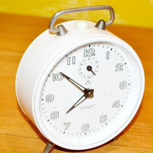 Könnte beinhalten: Weißer analoger Wecker mit schwarzen Zeigern und Zahlen. Das Zifferblatt hat ein kleineres, kreisförmiges Zifferblatt mit einem schwarzen Zeiger und Zahlen. Die Uhr ist mit "Peter" und "Made in Germany" beschriftet.