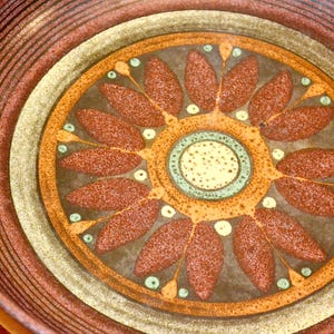 May include: A brown ceramic bowl with a concentric circle design. The center circle features a stylized flower with brown petals and a yellow center. The bowl has a rustic, earthy feel.