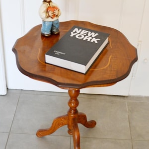 Puede incluir: Una pequeña mesa de pedestal de madera con borde festoneado, que presenta un libro negro titulado NEW YORK y una pequeña figura de cerámica de una persona con sombrero azul que sostiene un corazón rojo.