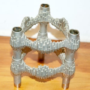 Peut inclure: Un bougeoir en métal argenté à trois pieds. Le bougeoir comporte trois plateformes circulaires avec des détails verticaux texturés. Chaque plateforme a une ouverture cylindrique pour une bougie. La pièce est en métal et repose sur une surface en bois.