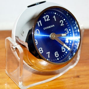 Könnte beinhalten: Ein silberner Wecker mit blauem Zifferblatt und weißen Zahlen. Die Uhr steht auf einem durchsichtigen Acryl-Ständer. Das Zifferblatt der Uhr zeigt "DUGENA".