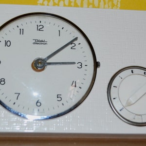 Może przedstawiać: Biały timer kuchenny z okrągłym cyferblatem i mniejszym okrągłym pokrętłem. Tarcza zegara ma czarne cyfry od 1 do 12 i napis "Diehl dilectron". Mniejsze pokrętło ma czarne oznaczenia i biały wskaźnik.