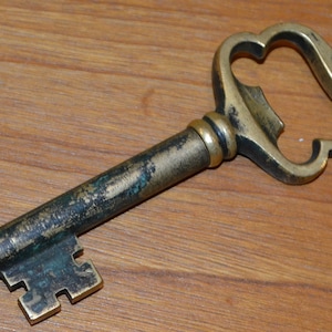 Può includere: Un apribottiglie vintage in ottone a forma di chiave. L'apribottiglie ha un grande anello decorato nella parte superiore e un piccolo design intricato a forma di buco della serratura nella parte inferiore.