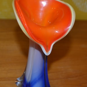 Może przedstawiać: Szklany wazon o unikalnym wzorze. Górna część wazonu ma kształt jaskrawo pomarańczowego i żółtego kwiatu. Dolna część wazonu jest niebieska i biała.