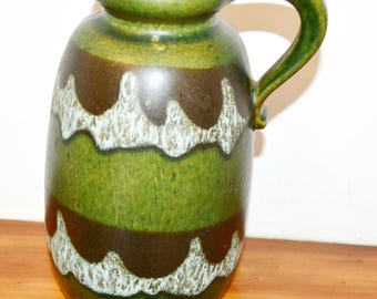 May include: A green and brown ceramic jug with a handle. The jug features a rounded body with horizontal bands of colour, including green, brown, and white wavy patterns. The handle is curved and attached to the side of the jug.