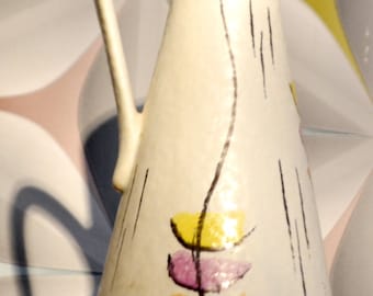Op de afbeelding: Een witte keramische kan met een handvat en een geometrisch ontwerp in geel, oranje en paars. De kan heeft een getextureerd oppervlak en een smalle hals.