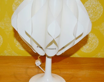 May include: White table lamp with a unique, pleated, white shade. The shade is made of multiple overlapping panels that create a geometric pattern. The lamp has a white base with a simple, elegant design.