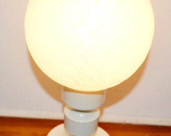 May include: A white, spherical table lamp with a glowing, round shade. The lamp has a cylindrical base with stacked, rounded sections. The lamp is plugged into a wall outlet. The lamp is on a wooden surface.