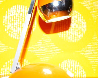May include: A vintage orange and chrome desk lamp with a round, swiveling shade. The lamp has a silver band around the middle of the shade.