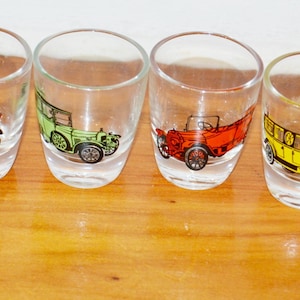 May include: Six clear shot glasses featuring vintage car designs in red, green, and yellow. The glasses are displayed on a wooden surface.