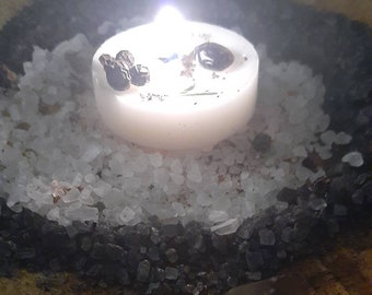 May include: A white candle with a black peppercorn and a clear crystal on a bed of white and black salt.