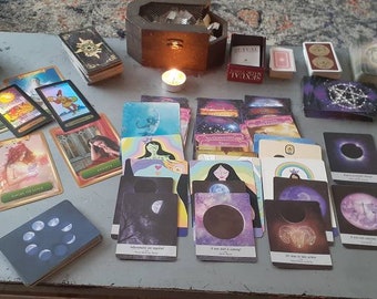 May include: A spread of tarot cards on a table, including a deck of oracle cards, a candle, and a wooden box. The cards are arranged in a circle and include images of moons, stars, and other celestial bodies.
