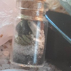 May include: A small glass bottle with a cork stopper, filled with dried herbs and crystals. The bottle is sitting on a white surface with other crystals.